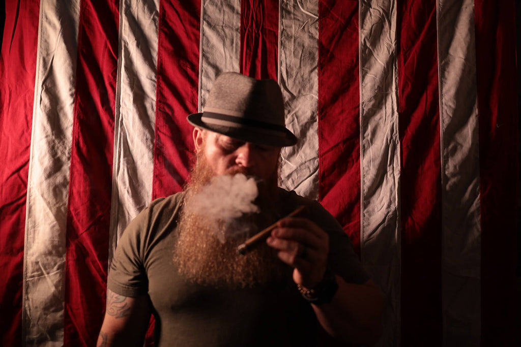 Mr. Luxurious standing in front of a flag smoking a cigar, Mens Beard care, oil, balm, butter wash Mr. Luxurious